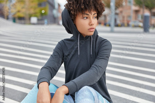 Horizontal shot of thoughtful swag African girl with pensive expression, dressed in hoodie and leggings, has rest on asphalt in sportsground, focused aside. Street lifestyle and youth concept