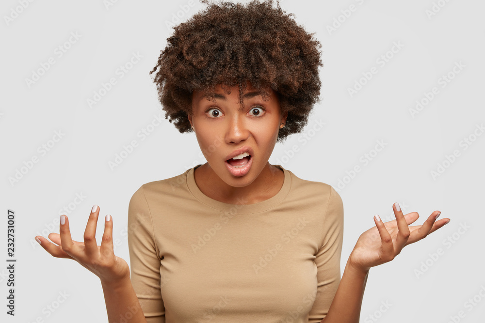 Photo of confused hesitant young woman has Afro curly hairstyle, shruggs shoulders, frowns face in bewilderment, dressed casually, cant understand something, isolated over white studio background Photo of confused hesitant young woman has Afro curly hairstyle, shruggs shoulders, frowns face in bewilderment, dressed casually, cant understand something, isolated over white studio background