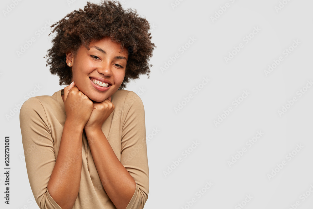 Studio shot of pleased emotive young Afro female model holds chin, has broad smile, perfect teeth, dressed casually, isolated over white studio wall, free space for your advertising content. Studio shot of pleased emotive young Afro female model holds chin, has broad smile, perfect teeth, dressed casually, isolated over white studio wall, free space for your advertising content.