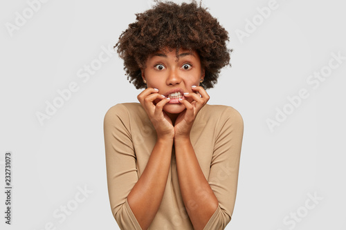 Stressful emotive young woman keeps hands near mouth, clenches teeth with anxious expression, has eyes full of fear, dressed in beige clothing, models against white studio wall. Omg concept.