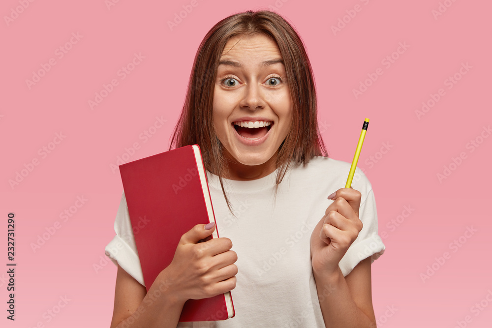 Rear view of joyful glad smiling woman with happy expression, holds red book and pencil in both hands, dressed in white t shirt, poses against pink background. People, emotions and education concept Rear view of joyful glad smiling woman with happy expression, holds red book and pencil in both hands, dressed in white t shirt, poses against pink background. People, emotions and education concept