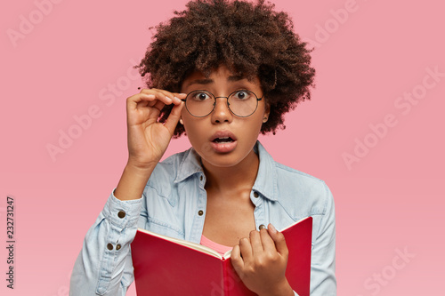Image of surprised black woman has Afro haircut, cant believe in bad exam result, wears round glasses, carries textbook, dressed in fashionable clothes, isolated over pink background. Omg concept