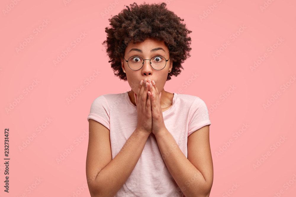 Picture of stupefied emotive black girl covers mouth with hands, stares at camera, exclaims in astonishment, wears round spectacles for good vision, hears terrible announcement from interlocutor Picture of stupefied emotive black girl covers mouth with hands, stares at camera, exclaims in astonishment, wears round spectacles for good vision, hears terrible announcement from interlocutor