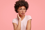 Isolated shot of tender young woman, has Afro bushy hairstyle, keeps hand on neck, wears round spectacles, dressed casually, isolated over pink background. People, youth and lifestyle concept.