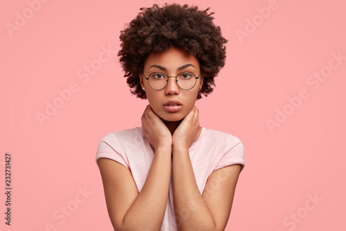 Headshot of serious beautiful African American woman keeps both hands on neck, wears round spectacles, looks directly at camera, isolated over pink background. People, beauty, lifestye concept.