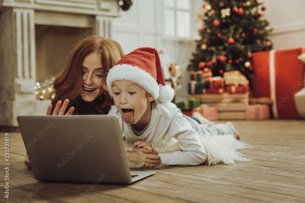 Online communication. Attractive child lying near his mother and demonstrating his tongue while staring at laptop Online communication. Attractive child lying near his mother and demonstrating his tongue while staring at laptop