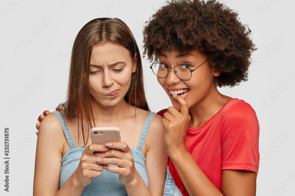 Studio shot of embarrased Caucasian woman looks at screen of mobile phone, texts messages, curious dark skinned girl embraces and looks with great interest, pose together against white background Studio shot of embarrased Caucasian woman looks at screen of mobile phone, texts messages, curious dark skinned girl embraces and looks with great interest, pose together against white background
