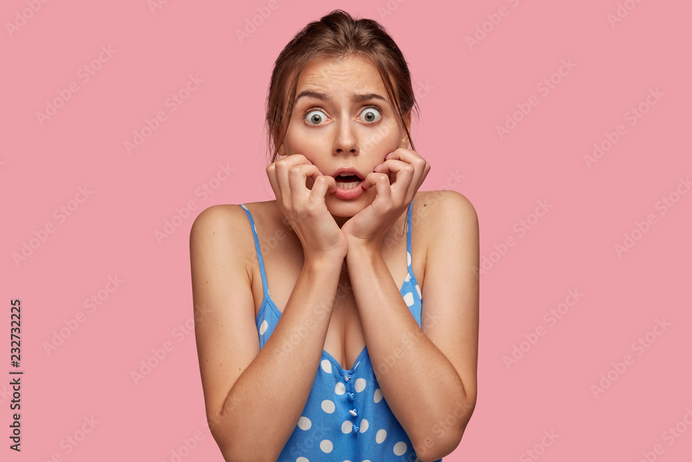 Scared beautiful woman looks with great fear, keeps both hands near mouth, being frightened by something, dressed in polka dot dress, isolated over pink background. Emotional girl afraids of nightmare Scared beautiful woman looks with great fear, keeps both hands near mouth, being frightened by something, dressed in polka dot dress, isolated over pink background. Emotional girl afraids of nightmare