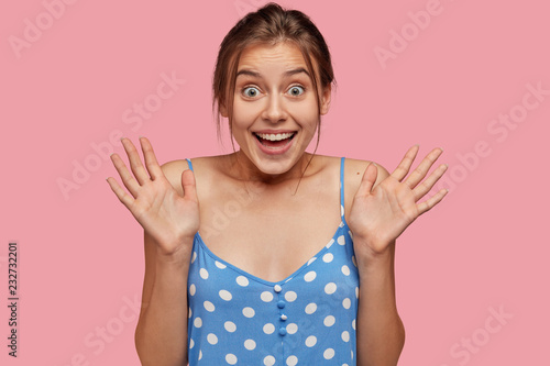 Photo of amazed pretty woman keeps hands in front, reacts on sudden surprising positive news, has eyes popped out from wonder, dressed in casual outfit, isolated over pink background. Emotions concept