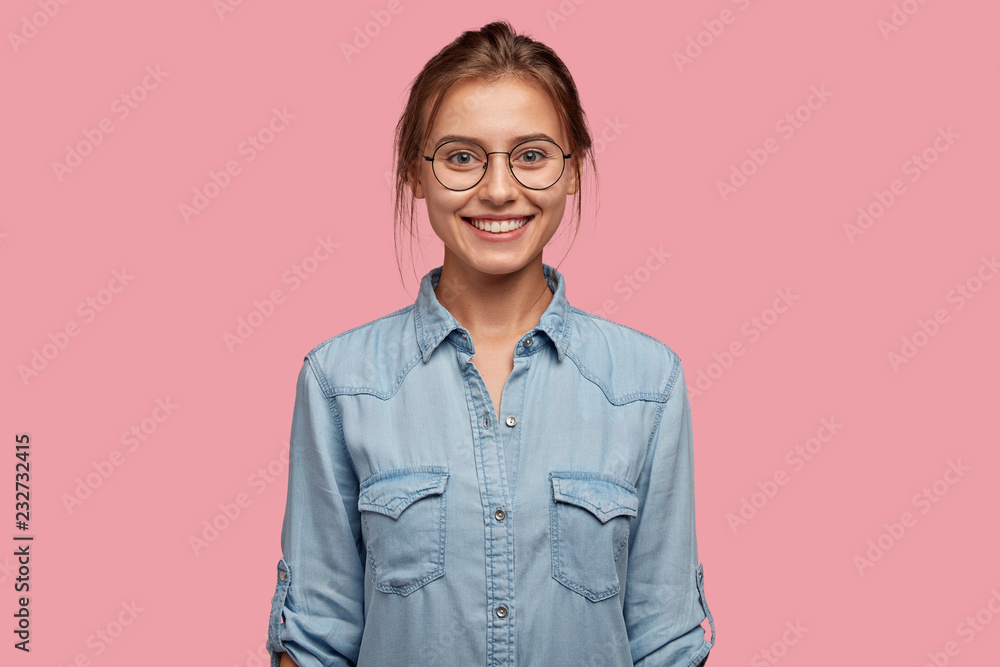Studio shot of smiling glad woman has appealing appearance, smiles broadly, dressed in denim shirt, poses against pink background expresses positive emotions likes what she sees, rejoices new purchase