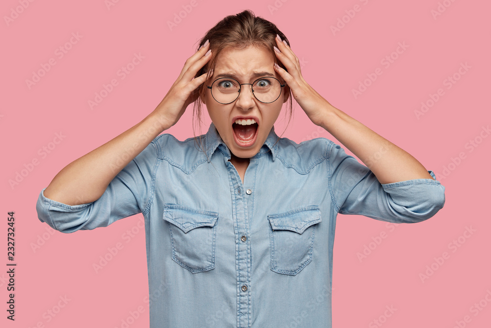 Emoitional young girl cries and screams agrily, keeps hands on head, opens mouth widely, dressed in denim shirt, expresses hate and rage, isolated over pink background. Negative human emotions concept Emoitional young girl cries and screams agrily, keeps hands on head, opens mouth widely, dressed in denim shirt, expresses hate and rage, isolated over pink background. Negative human emotions concept