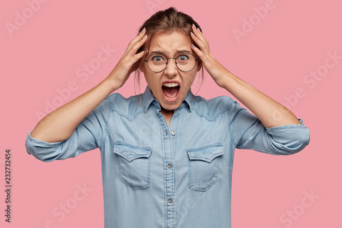 Emoitional young girl cries and screams agrily, keeps hands on head, opens mouth widely, dressed in denim shirt, expresses hate and rage, isolated over pink background. Negative human emotions concept