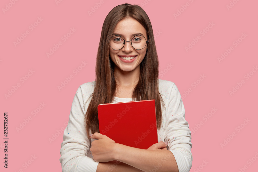 Waist up shot of pretty girl smiles pleasantly, carries red textbook closely, wears white casual jamper, models against pink background. Headshot of Caucasian lady student happy after having classes Waist up shot of pretty girl smiles pleasantly, carries red textbook closely, wears white casual jamper, models against pink background. Headshot of Caucasian lady student happy after having classes
