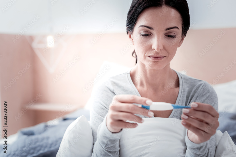 Checking temperature. Close up of young woman feeling sad while sitting on the bed and looking at thermometer Checking temperature. Close up of young woman feeling sad while sitting on the bed and looking at thermometer