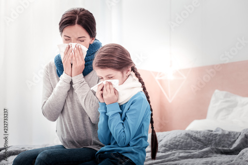 Falling ill together. Portrait of young mother and little daughter sitting on the bed while using handkerchiefs and blowing their noses