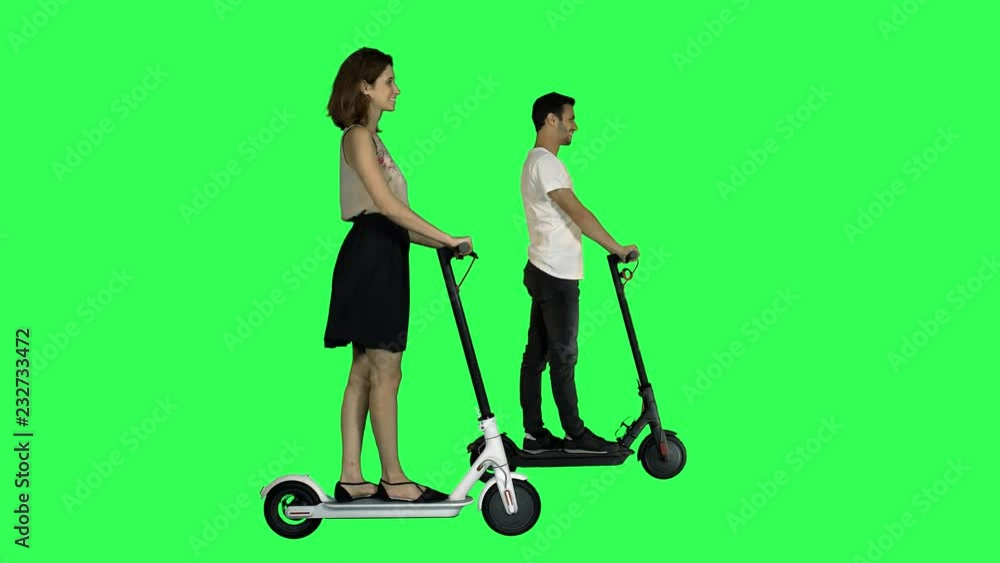 Happy couple riding scooters together, looking at each other, smiling, over a green screen, woman at the front. Happy couple riding scooters together, looking at each other, smiling, over a green screen, woman at the front.
