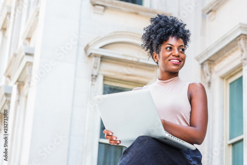 Way to Success. Young African American woman with afro hairstyle wearing sleeveless light color top, sitting by vintage office building in New York, working on laptop computer, looking up, smiling..