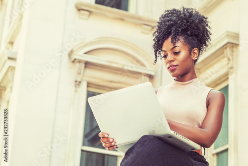 Way to Success. Young African American woman with afro hairstyle wearing sleeveless light color top, sitting by vintage office building in New York, looking down, reading, working on laptop computer