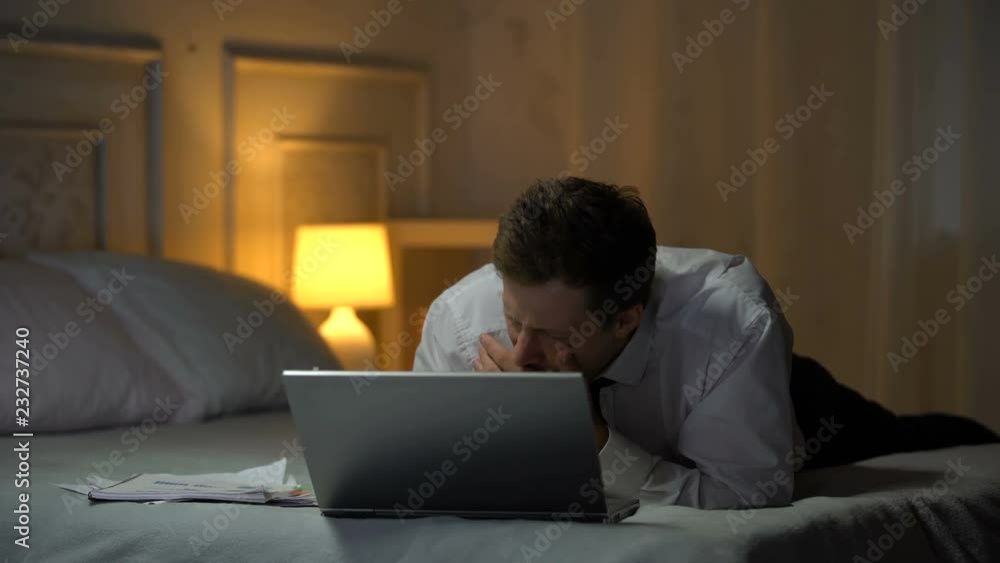 Exhausted engineer lying on bed, working on laptop and yawing, reading mail Exhausted engineer lying on bed, working on laptop and yawing, reading mail