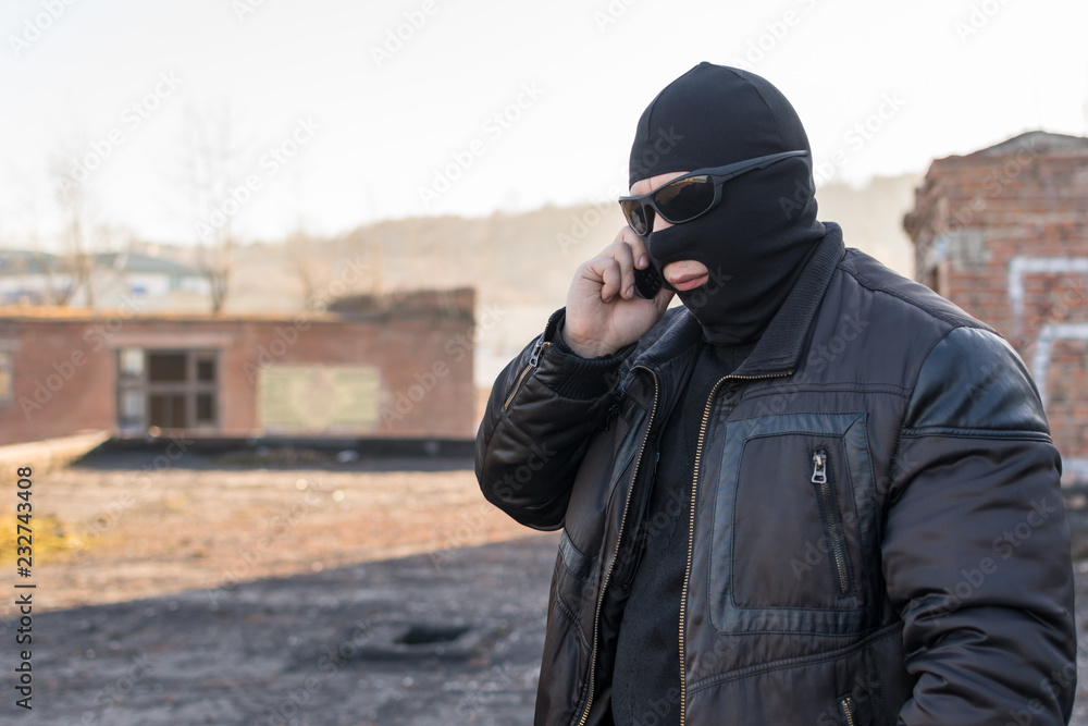 a man, a terrorist, a bandit in a black leather jacket and a mask talking on the phone on the street near an abandoned building a man, a terrorist, a bandit in a black leather jacket and a mask talking on the phone on the street near an abandoned building
