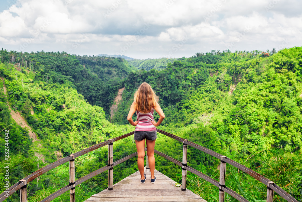 Family vacation lifestyle. Young woman stand on edge of overhanging bridge on high cliff. Happy girl looking at stunning tropical jungle view. Tukad Melangit is popular travel destination in Bali.