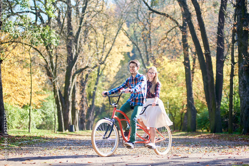 Young tourist couple, handsome bearded man and attractive blond long-haired woman in glasses cycling together tandem double red bike by sunny alley with golden leaves on tall trees background