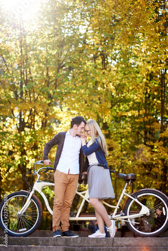Young happy romantic couple in love, bearded guy and attractive girl standing close together at tandem double bicycle in autumn park or forest on sunny bright golden yellow foliage bokeh background. © anatoliy_gleb