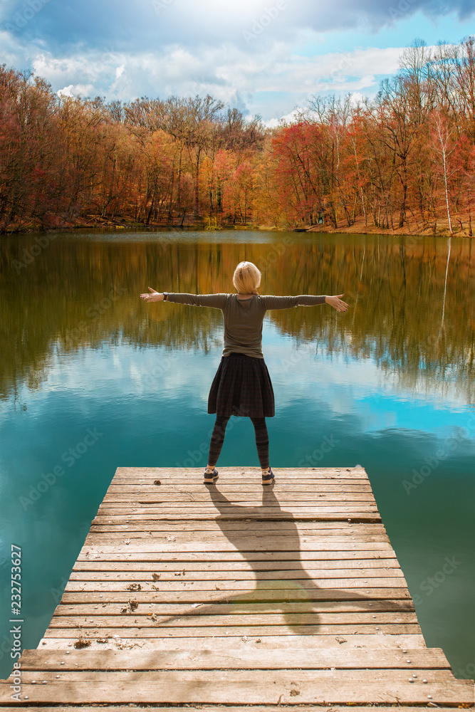 Woman standing on the edge of wooden dock, with her arms raised up, looking at water and colorful autumnal forest