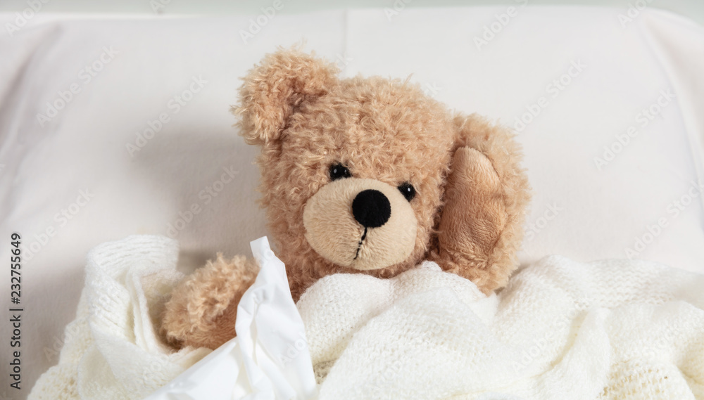 Cold, flu or allergy. Cute teddy in bed, covered with a warm blanket, holding a tissue