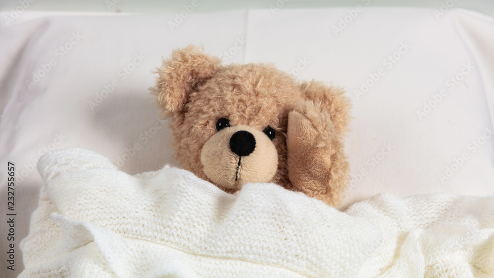 Headache, insomnia. Cute teddy in bed, covered with a warm blanket, holding his head