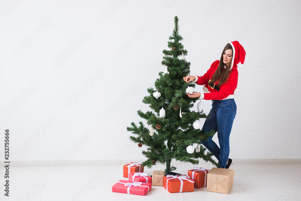 Winter holidays, xmas and people concept - Portrait of beautiful young woman in Santa hat decorating Christmas tree at home in white background with copy space Winter holidays, xmas and people concept - Portrait of beautiful young woman in Santa hat decorating Christmas tree at home in white background with copy space