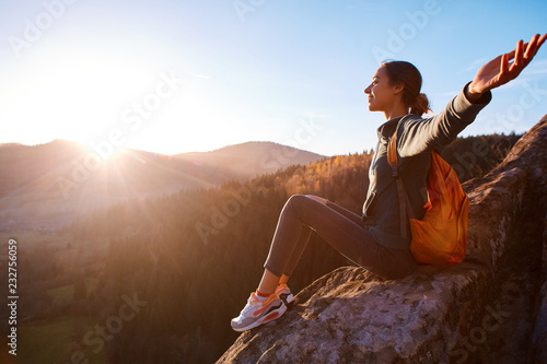 woman hiker with backpack sits on edge of cliff against background of sunrise. Woman is meditation and greeting a sun