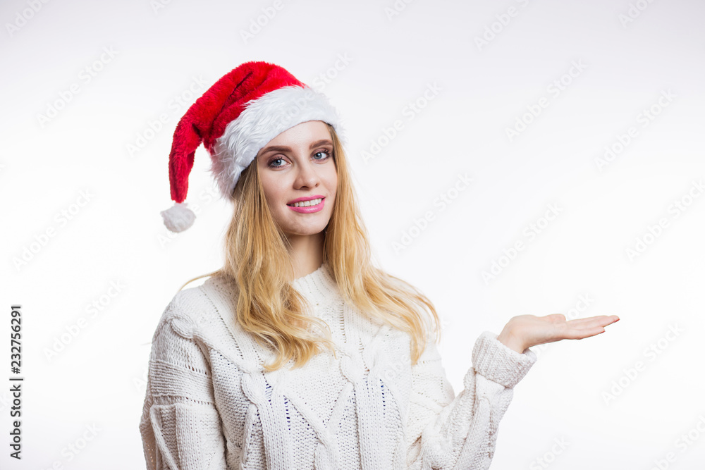 Christmas portrait of attractive young blonde woman in beige warm sweater and red Santa hat showing your product Christmas portrait of attractive young blonde woman in beige warm sweater and red Santa hat showing your product