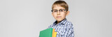 A charming boy with a vkletchatoy shirt and light jeans stands on a gray background. The boy holds in his hands a multicolored folder with documents