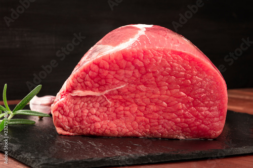 A photo of eye round beef, a raw cut, with rosemary, garlic, and pepper, on a black background with copy space photo