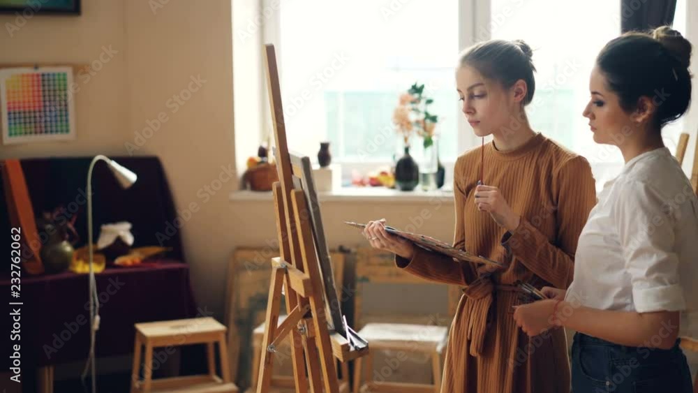 Cheerful art student is working at picture painting with brush on canvas while her teacher is checking her work and helping her. Girl is smiling enjoying class. Cheerful art student is working at picture painting with brush on canvas while her teacher is checking her work and helping her. Girl is smiling enjoying class.