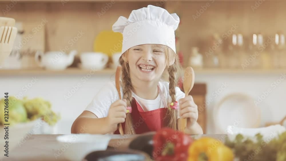 Portrait of a cute little girl in the kitchen dressed as a professional cook looking into the camera and smiling. She holds a fork and a spoon in her hands and have fun. Portrait of a cute little girl in the kitchen dressed as a professional cook looking into the camera and smiling. She holds a fork and a spoon in her hands and have fun.