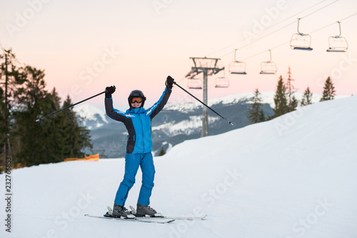 Girl enjoying ski holiday standing on the snowy mountain and raised her hands up. Woman at ski resort wearing helmet, blue ski suit and goggles.