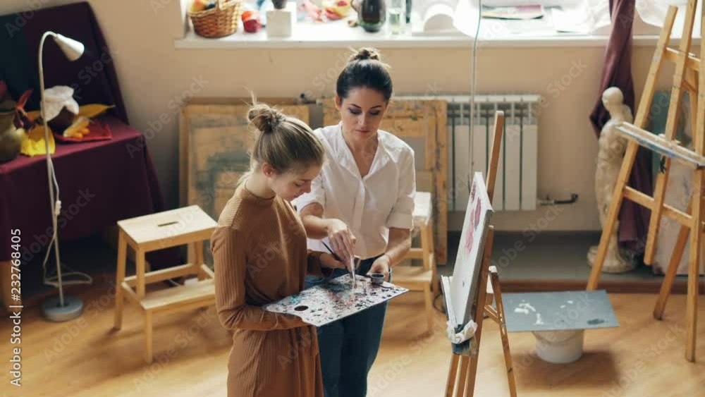 Good-looking young woman is holding palette and painting flowers under guidance of helpful teacher standing together in workroom of art school and talking. Good-looking young woman is holding palette and painting flowers under guidance of helpful teacher standing together in workroom of art school and talking.
