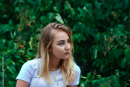 Cute charming blonde in a white blouse looks into the distance on a background of green vegetation.