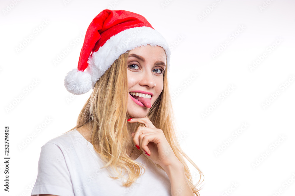 Funny cheerful smiling blonde woman wears a red Santa hat and shows her tongue to the camera on a white background Funny cheerful smiling blonde woman wears a red Santa hat and shows her tongue to the camera on a white background