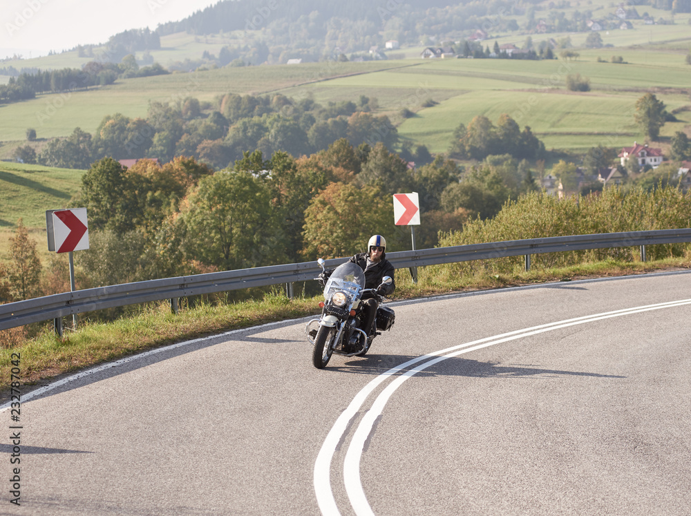 Bearded motorcyclist in helmet, sunglasses and black leather clothing driving bike along sharp turn of empty road on bright summer day on misty background of rural landscape and distant green hills.