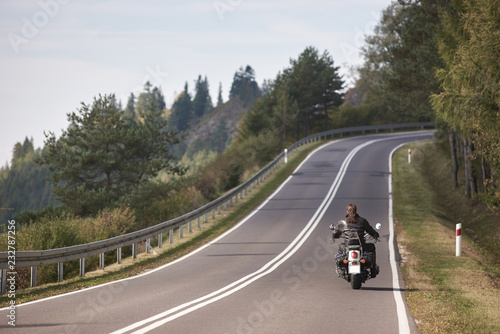Back view of motorcyclist in black leather outfit with long hair riding cruiser motorbike up twisted empty asphalt road on bright sunny summer day by green forest trees.