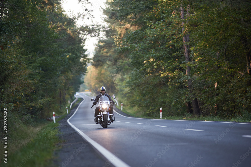 Bearded biker in sunglasses, helmet and black leather clothing riding modern powerful high-speed motorcycle along asphalt road winding among tall green trees.