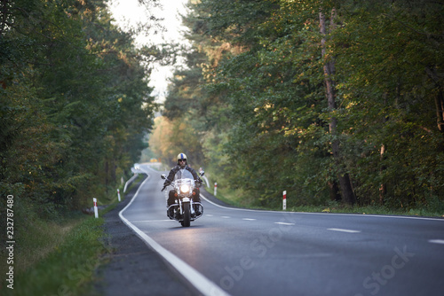 Bearded biker in sunglasses, helmet and black leather clothing riding modern powerful high-speed motorcycle along asphalt road winding among tall green trees.