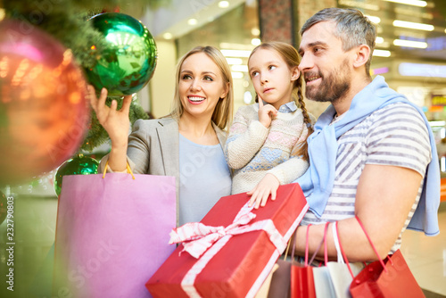 Couple with little daughter looking at baubles on Christmas tree while shopping together