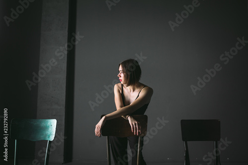 The social theme female loneliness pain suffering. Abastration male violence family. A young beautiful Caucasian woman in black clothes sitting on three chairs in large dark room. Srach and despair photo