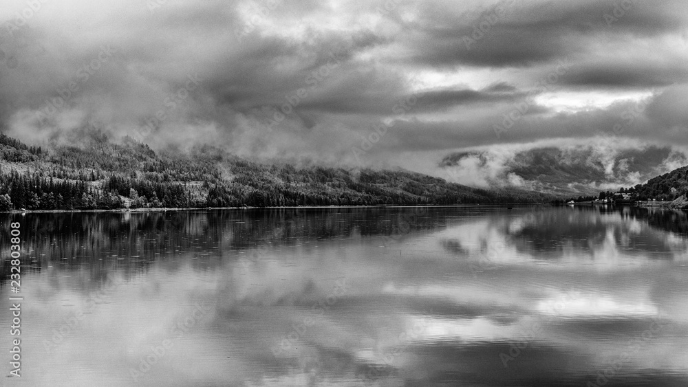Fototapeta premium Beautiful reflection in the Tyrifjorden (Lake Tyri) after a rainy day in Norway. Dramatic sky, fog, trees and mountains are perfectly mirrored in the lake. Black and white picture