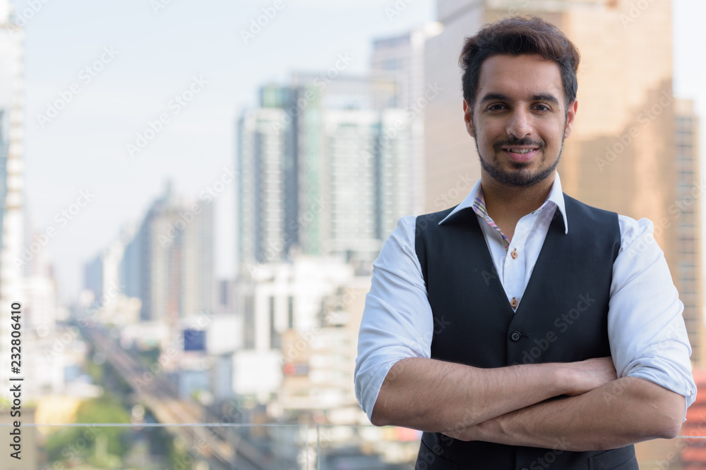 Young happy Indian man smiling with arms crossed at rooftop in B