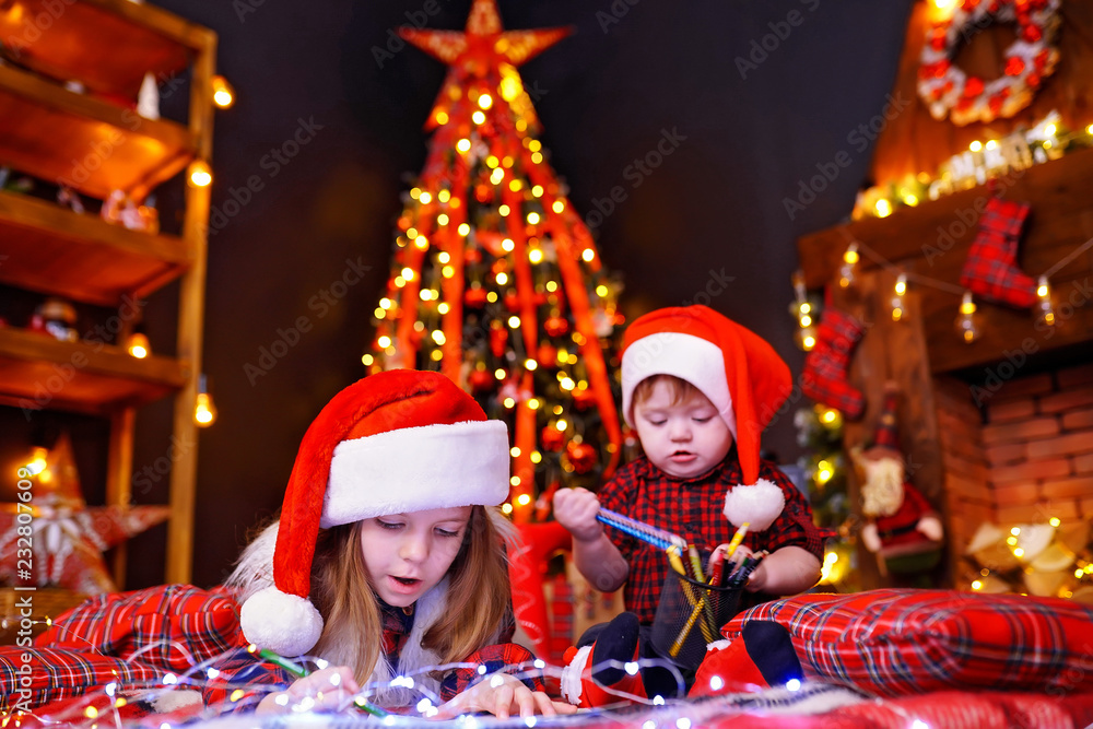 Charming girl in Santa hat lying on the floor and writes letter, draws with pencil and her little brother who interferes with her, plays with pencils in warm room with garlands, lights